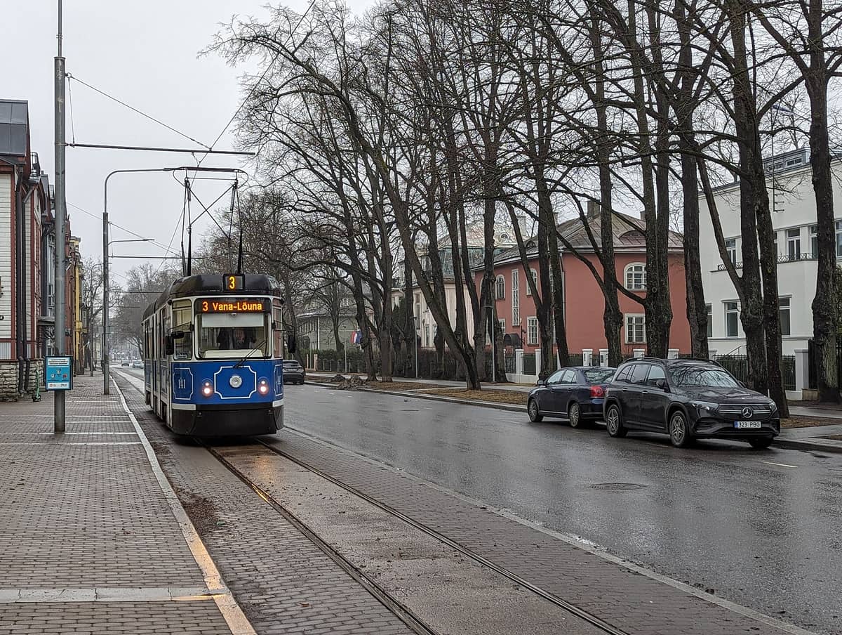 Trem biru dan putih Tallinn di jalur 3 yang melaju di sepanjang jalan kota yang dipenuhi pepohonan dan mobil parkir.