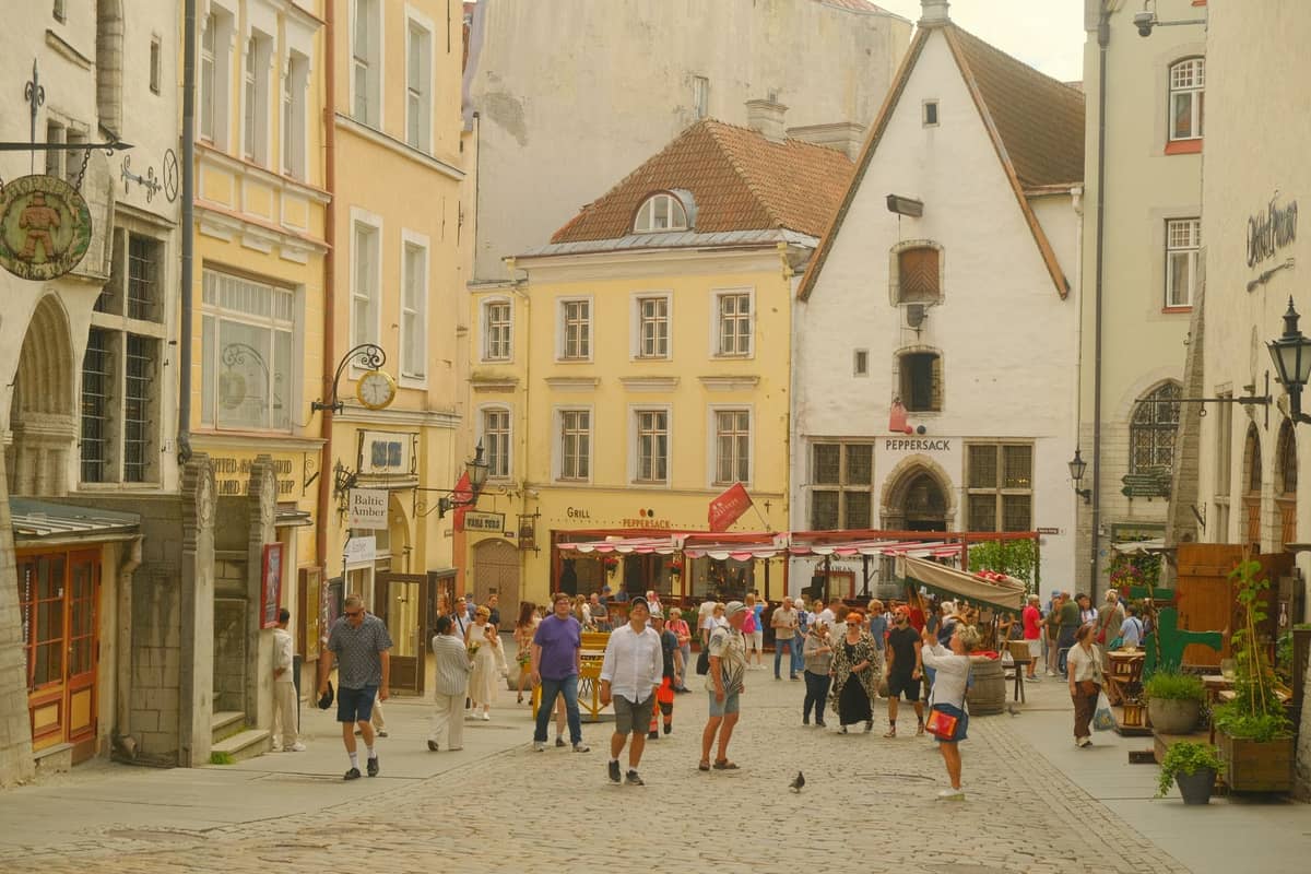 Kerumunan orang berjalan di sepanjang jalan berbatu yang dilapisi bangunan-bangunan abad pertengahan berwarna-warni di Kota Tua Tallinn.