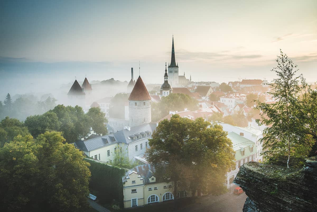 Pemandangan lanskap Tallinn yang menampilkan Kota Tua abad pertengahan di atas bukit batu kapur dengan menara-menara bersejarah, menara gereja, dan tata kota yang terlihat di bawah kabut pagi.