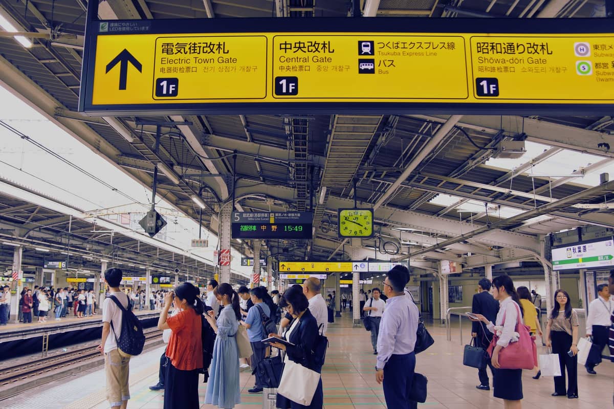 Crowds of people waiting on the platform of a busy Tokyo train station, with yellow overhead signs showing directions, gates and train lines.