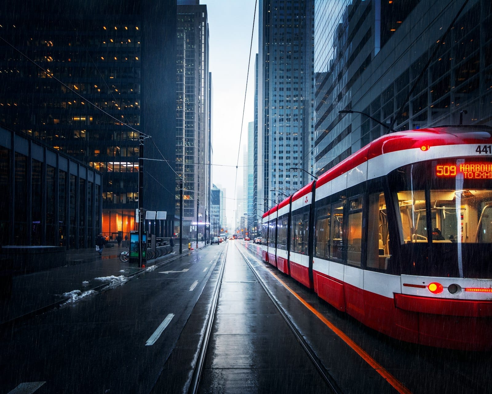 Um bonde moderno vermelho e branco de Toronto percorre uma rua do centro em um dia chuvoso, ladeada por arranha-céus de vidro.