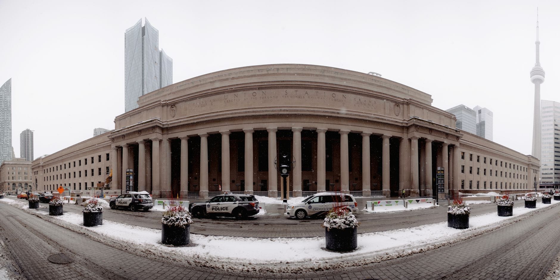 Vista em grande angular do exterior da Union Station de Toronto no inverno, mostrando suas grandes colunas, neve no chão e a CN Tower nas proximidades.