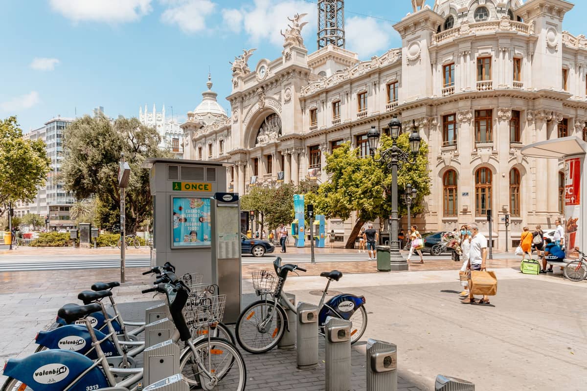 Stazione di bike sharing Valenbisi con persone che camminano davanti all'ornato palazzo delle Poste nel centro di Valencia in una giornata di sole.
