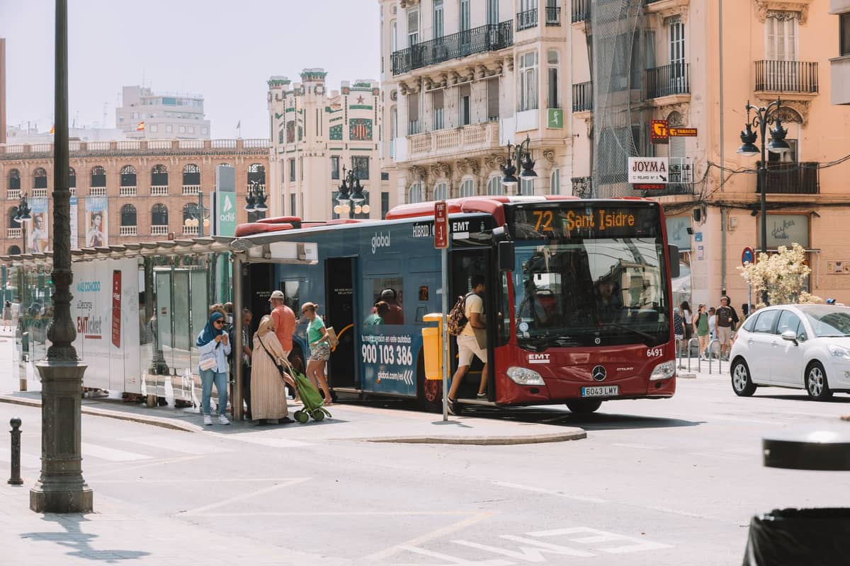 Autobús rojo de la EMT Valencia en una parada central, personas subiendo y bajando, con edificios históricos de la ciudad al fondo.