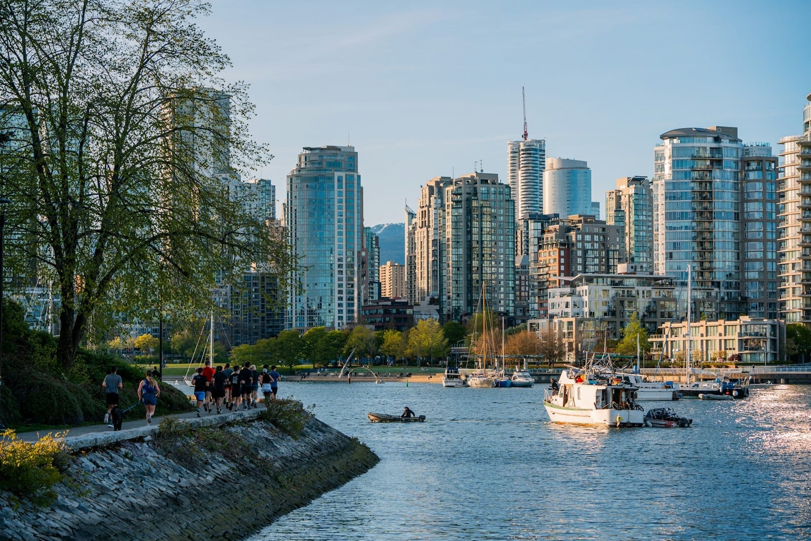 Pessoas caminham e pedalam pela orla marítima de Vancouver à beira-d'água, com barcos e arranha-céus do centro ao fundo sob um céu azul.