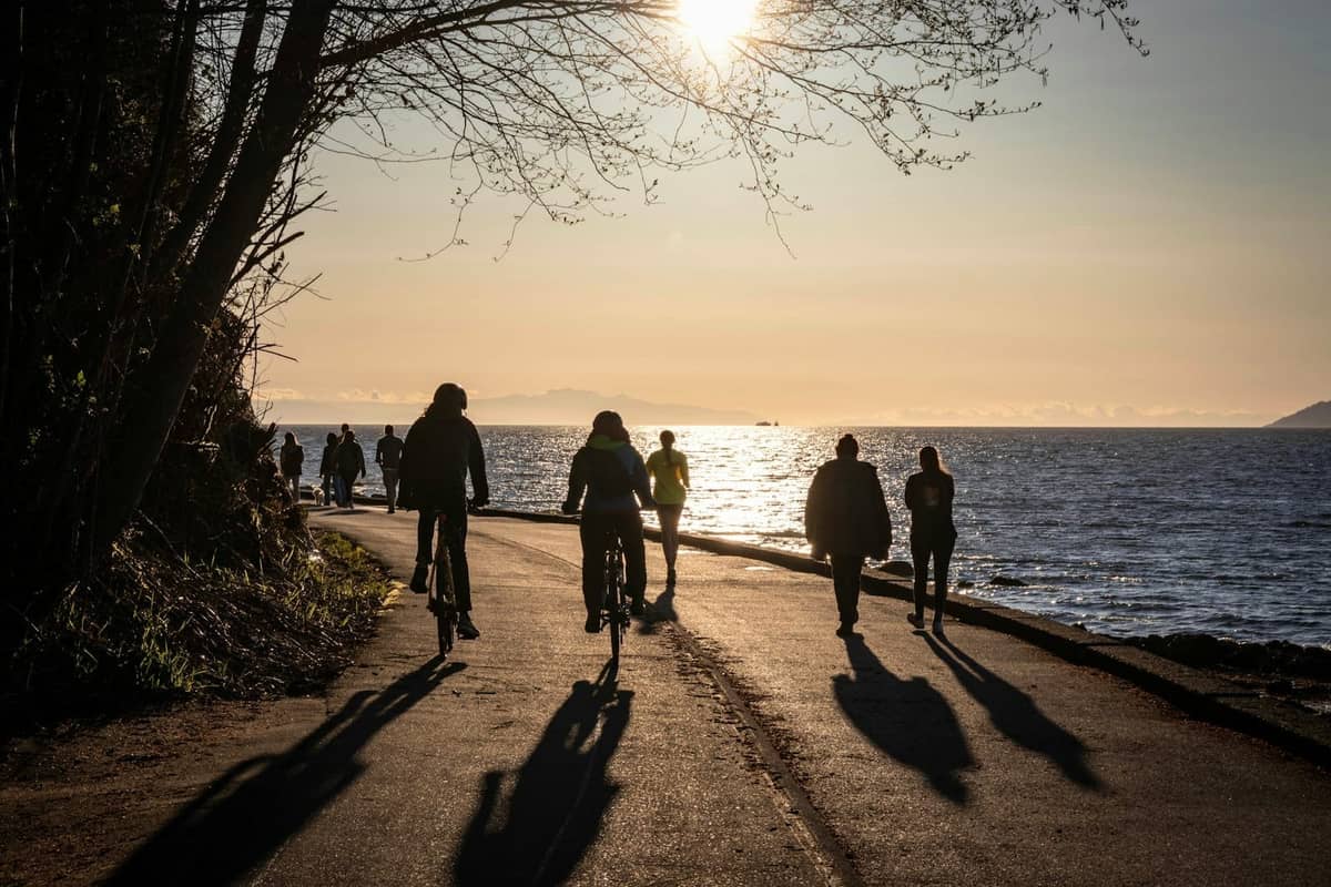 Menschen, die bei Sonnenuntergang entlang Vancouvers malerischem Seawall-Pfad spazieren und Fahrrad fahren, mit sichtbarem Ozean und Bäumen.