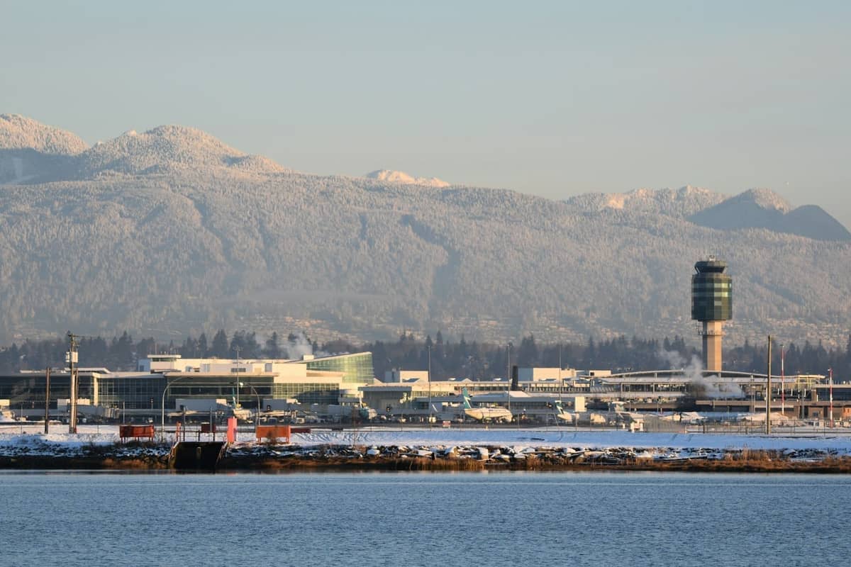Weitwinkelansicht des Terminals des Vancouver International Airport (YVR) mit Kontrollturm und schneebedeckten Bergen im Hintergrund, vom Wasser aus gesehen.