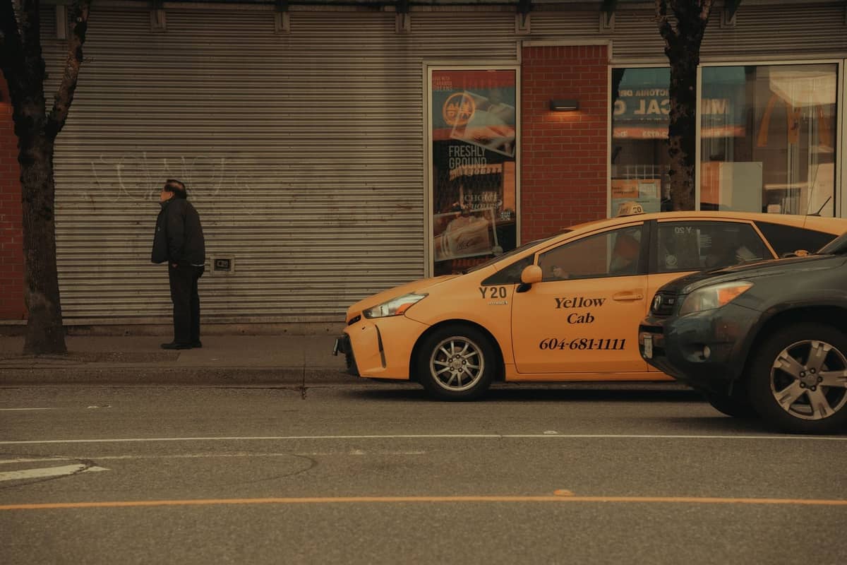 Ein Yellow Cab-Taxi parkt tagsüber auf einer Straße in Vancouver, in der Nähe ein Fußgänger, Geschäfte und Bäume entlang des Gehwegs.