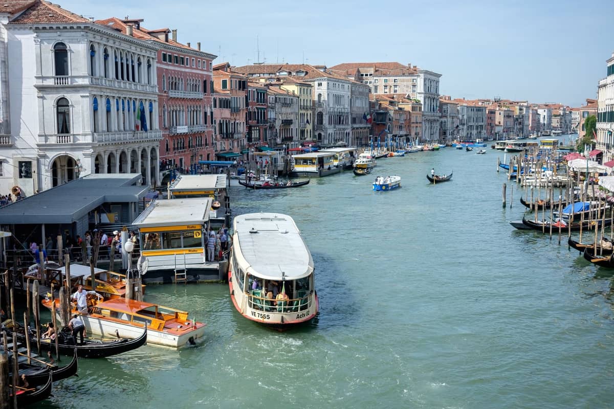 Belebter Canal Grande in Venedig mit Wasserbussen, Gondeln und Booten zwischen historischen Gebäuden an der Uferpromenade an einem sonnigen Tag.