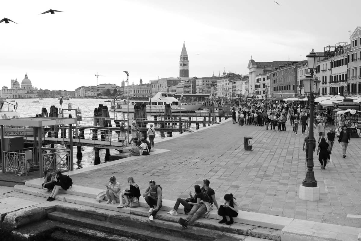 Menschenmengen schlendern und versammeln sich entlang der belebten Uferpromenade von Venedig mit angedockten Booten und Stadtsehenswürdigkeiten im Hintergrund.