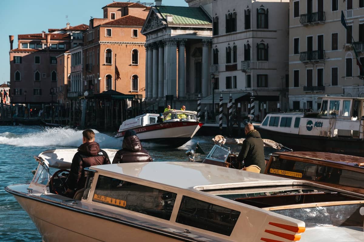 Mehrere Wassertaxis mit Passagieren, die auf einem belebten Kanal in Venedig fahren, umgeben von klassischen venezianischen Gebäuden bei klarem Tageslicht.