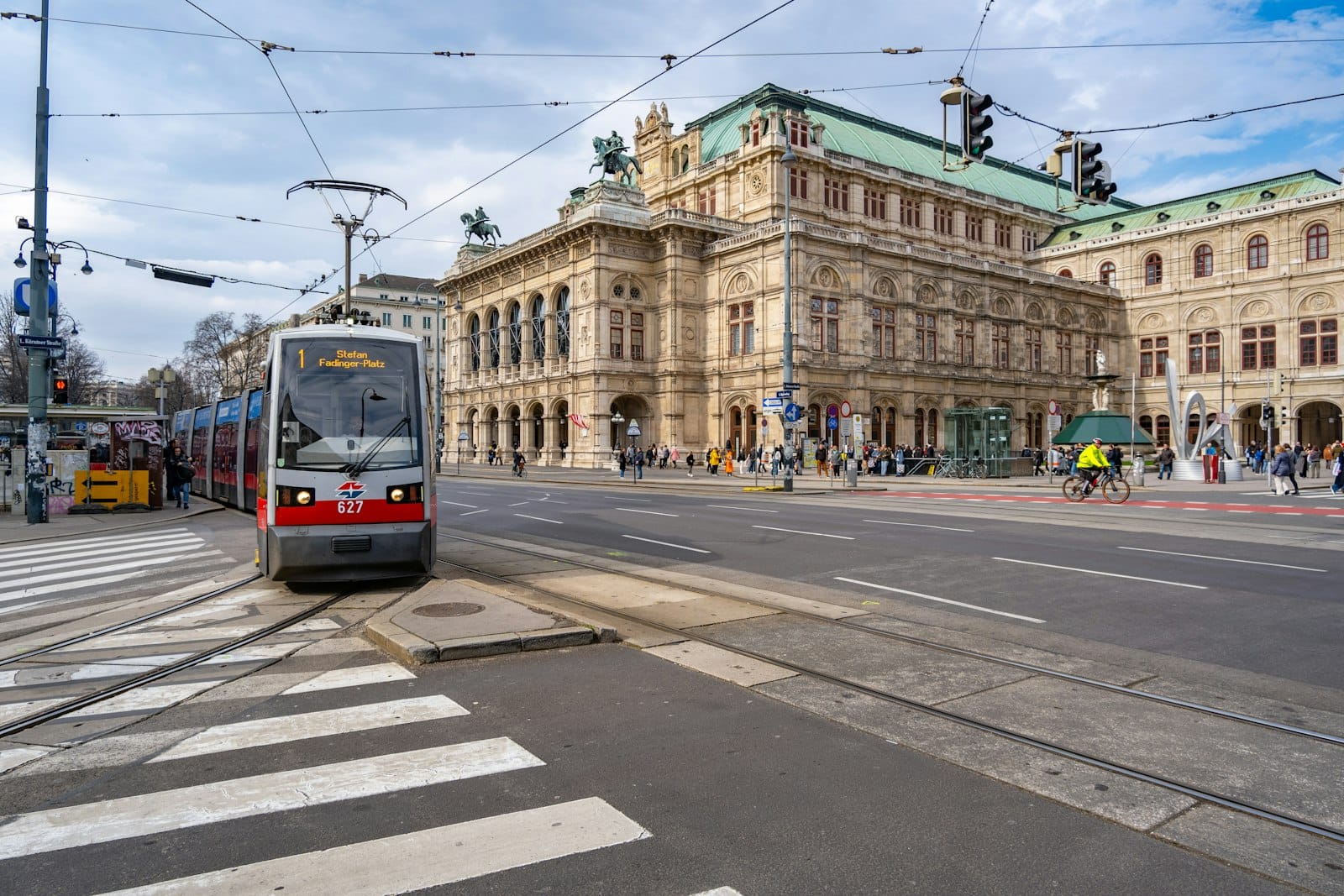Eine moderne Wiener Straßenbahn fährt vor der historischen Wiener Staatsoper vorbei, während Menschen, Autos und Fahrradfahrer die belebte Stadtszene teilen.