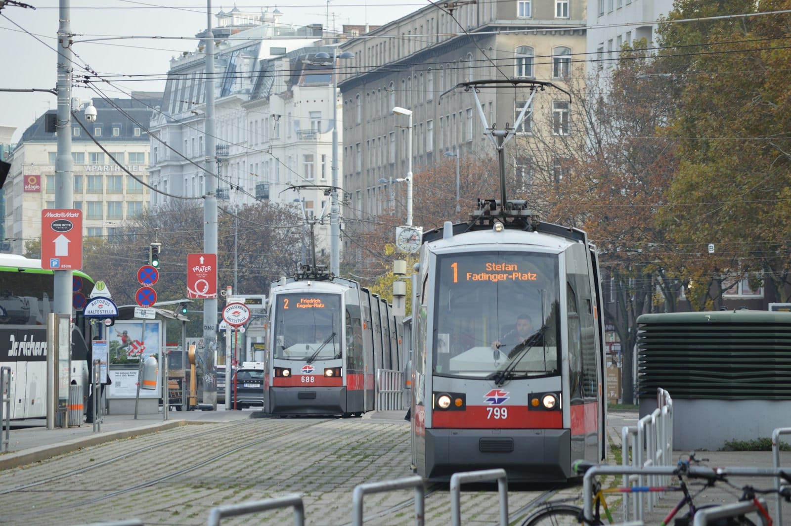 Due moderni tram della Wiener Linien su binari cittadini con edifici di Vienna, una fermata del tram e persone sullo sfondo.