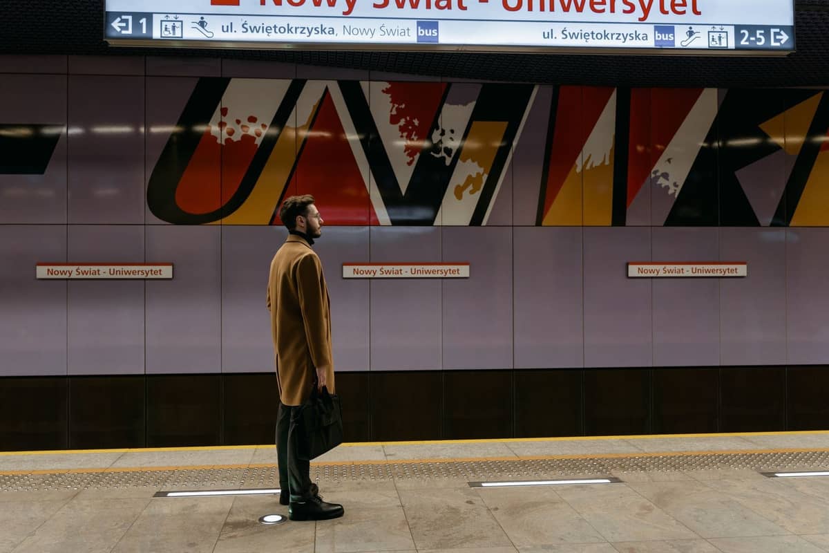 Pria menunggu di peron stasiun metro modern Nowy Świat - Uniwersytet di Warsawa dengan papan petunjuk yang terlihat.