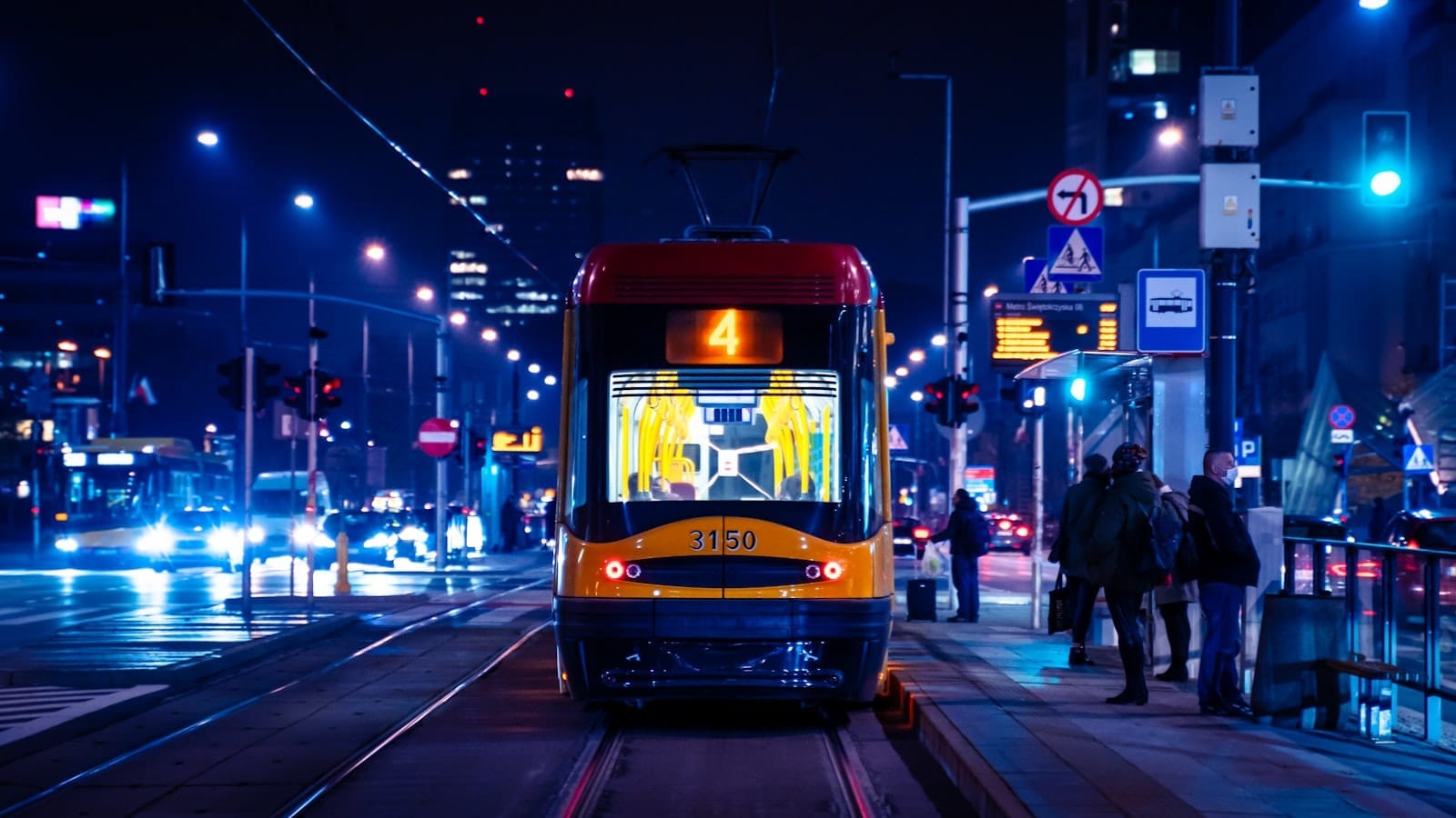 Bonde moderno de Varsóvia à noite no centro da cidade, com postes iluminados, pessoas esperando na parada e prédios urbanos ao fundo.