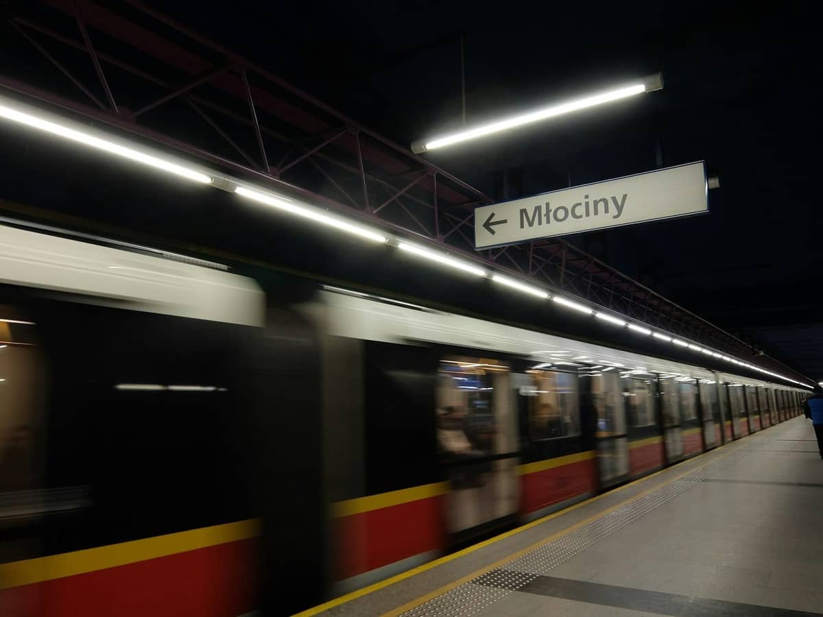 Peron metro Warsawa dengan kereta yang sedang bergerak dan tanda stasiun Mlociny di atas, menampilkan sistem transportasi kota modern.