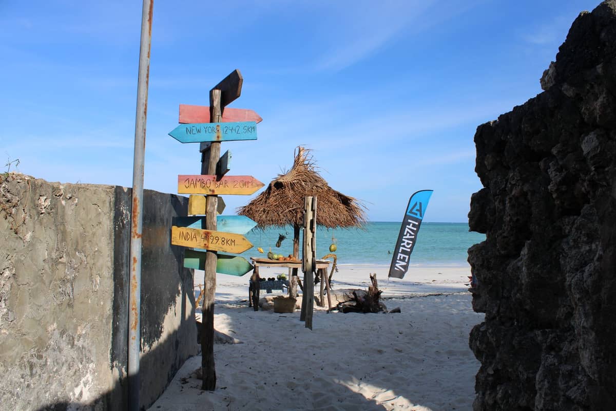 Colorful wooden signposts on a sandy path leading to a thatched hut and turquoise beach in Zanzibar, suggesting directions between beach areas.