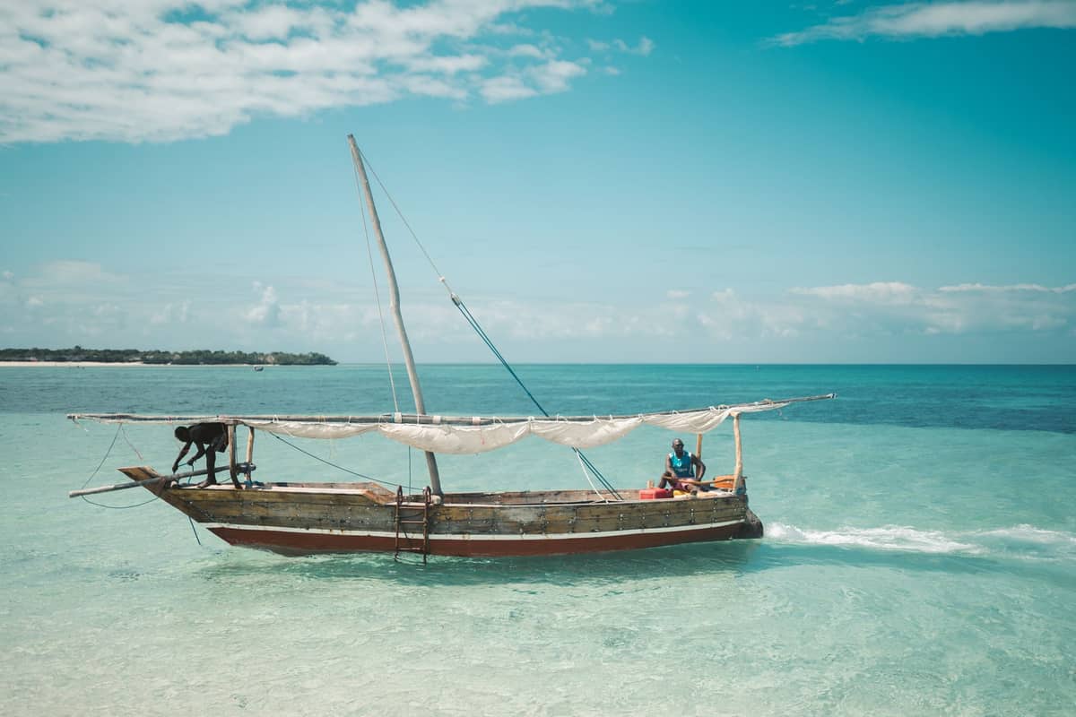 A traditional wooden dhow boat with two people onboard sails across clear turquoise water under a blue sky in Zanzibar.