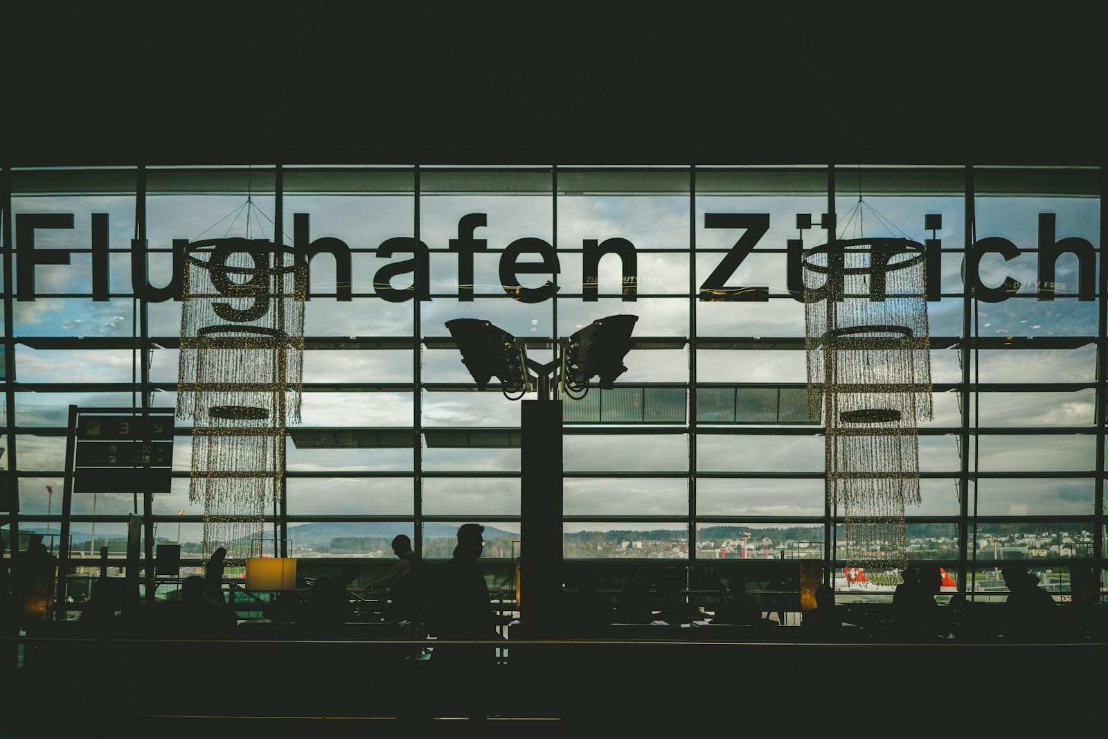หน้าต่างกระจกขนาดใหญ่ที่ Flughafen Zürich พร้อมข้อความ Flughafen Zürich เงาของนักเดินทาง และโครงสร้างสนามบินที่มองเห็นได้จากด้านนอก