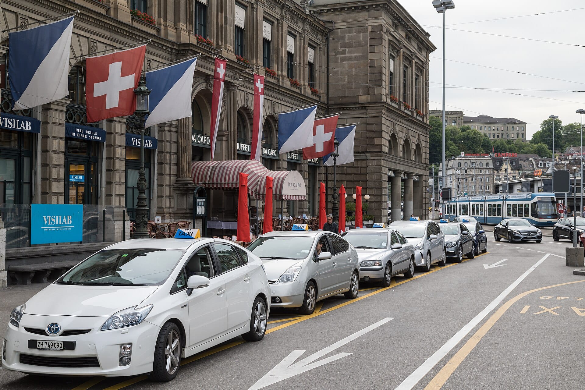 แถวแท็กซี่อย่างเป็นทางการจอดรอบริเวณนอกสถานีรถไฟหลักของ Zurich โดยมีธงชาติสวิตเซอร์แลนด์และรถรางให้เห็นในพื้นหลัง