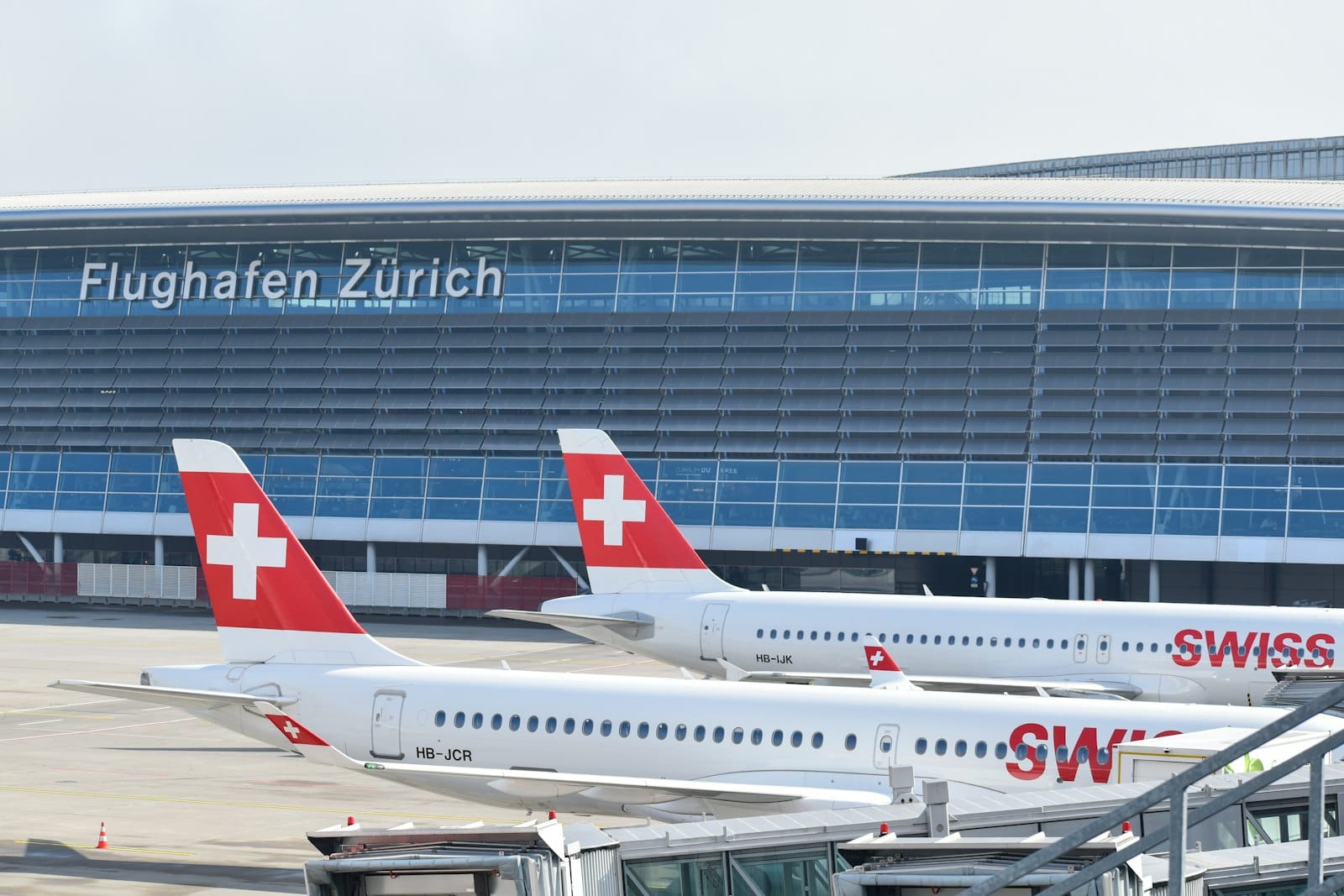 อาคารสนามบินซูริคพร้อมป้าย 'Flughafen Zürich' และเครื่องบิน SWISS จอดริมเกทในวันที่อากาศแจ่มใส