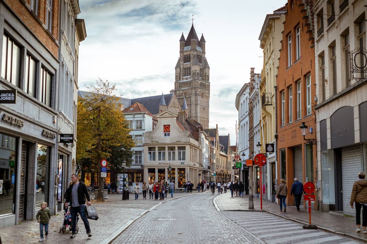 Pedestrian-friendly street in Bruges with people walking, cyclists, a bus lane, and the Belfry tower visible in the background.