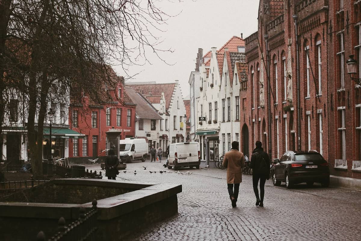 Pedestrians walk on a cobblestone street lined with historic brick and stepped-gable buildings in Bruges on a cloudy day.