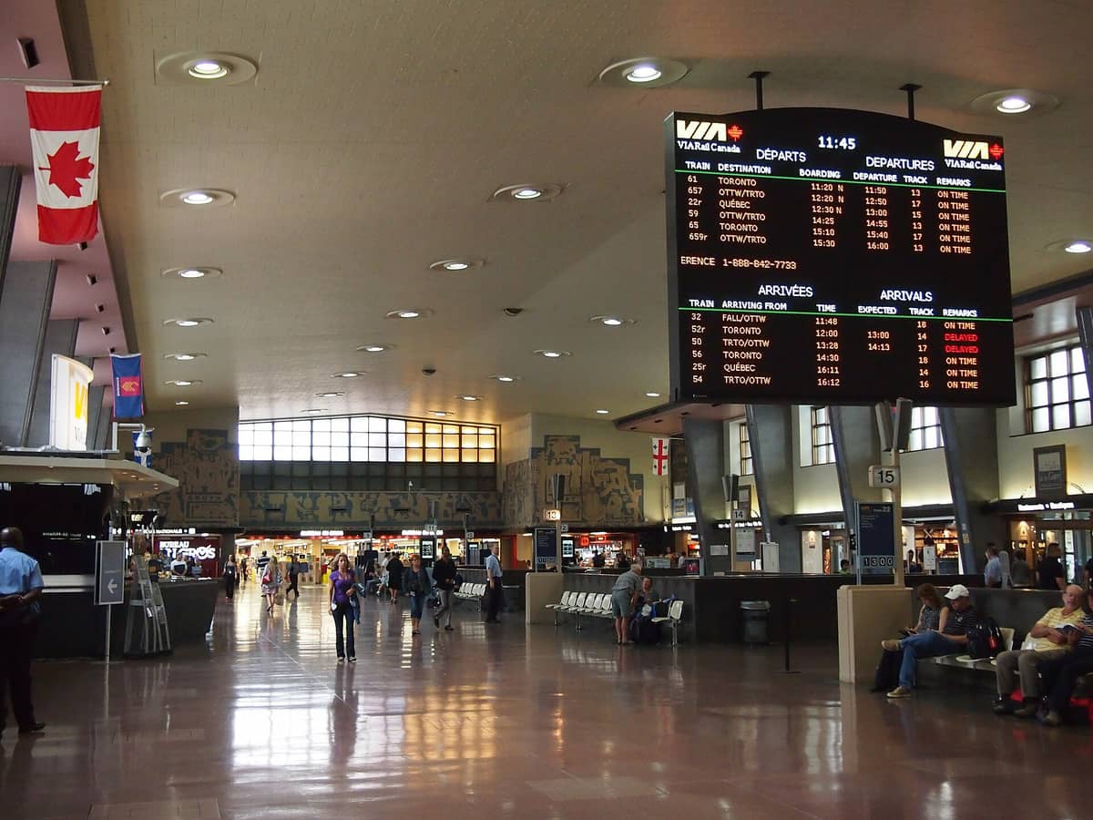 Interior da Estação Central de Montreal com um painel de partidas e chegadas da VIA Rail Canada, pessoas esperando, bandeira canadense e lojas visíveis.