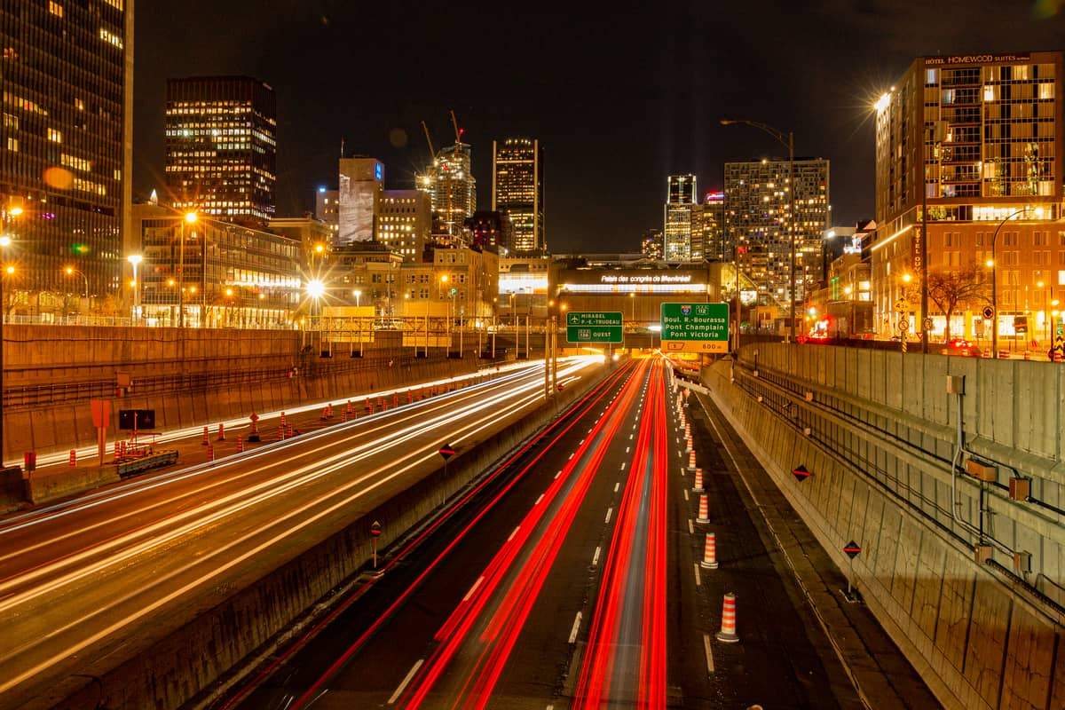 Vista noturna do centro de Montreal com arranha-céus, tráfego na autoestrada e placas de sinalização verdes elevadas para rotas urbanas.
