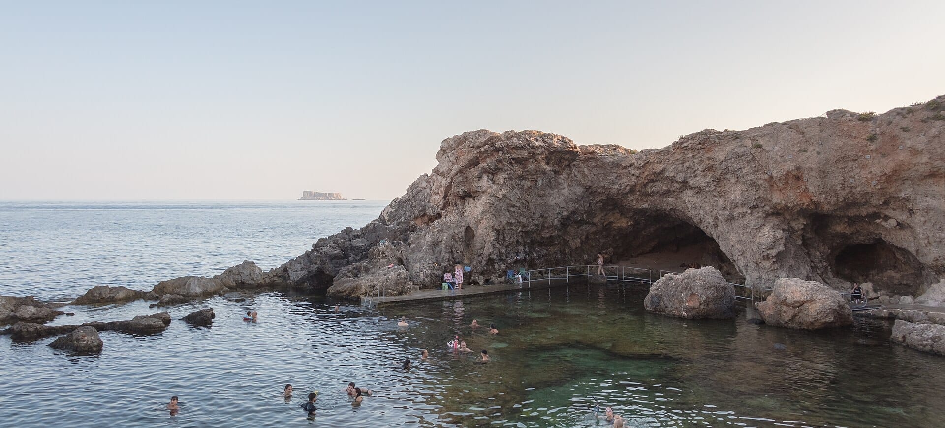 Acqua di mare limpida con nuotatori vicino all'ingresso roccioso della grotta Għar Lapsi, circondata da scogliere frastagliate sulla costa meridionale di Malta.