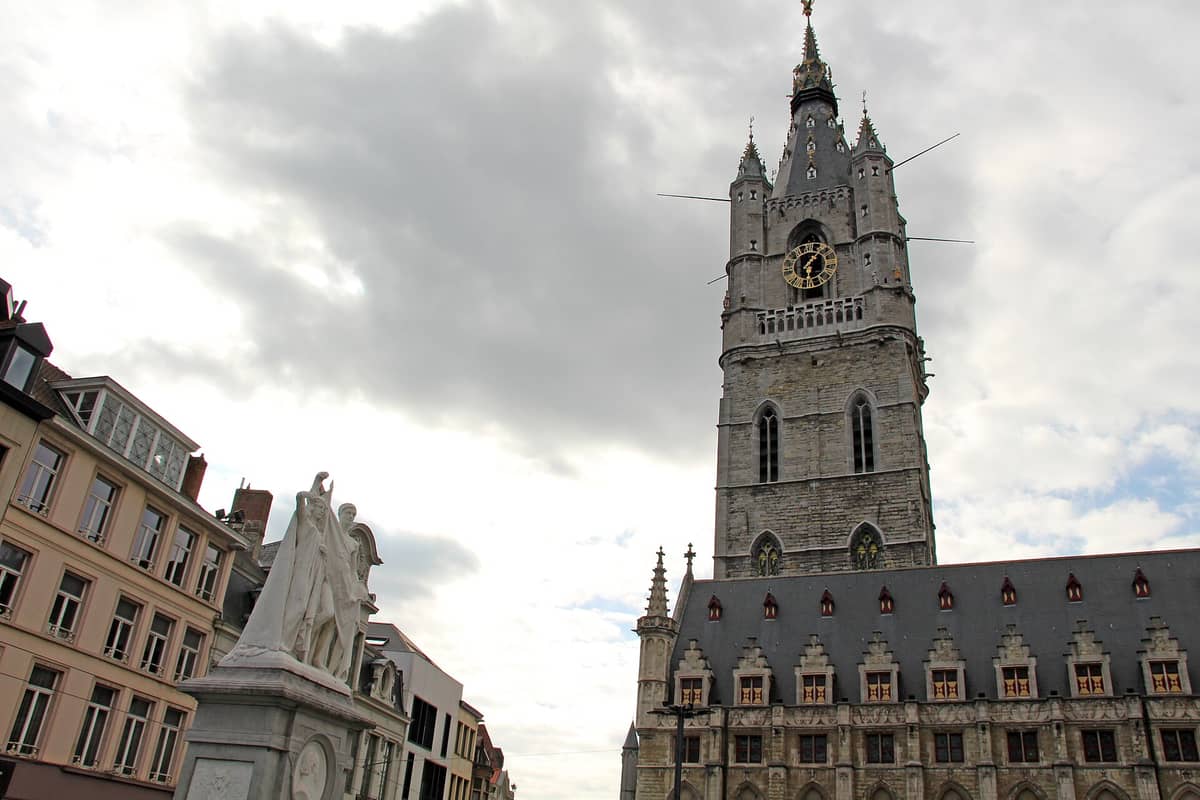 Wide view of the Ghent Belfry (Belfort van Gent) rising above a row of historic buildings and a statue in Sint-Baafsplein under a cloudy sky.