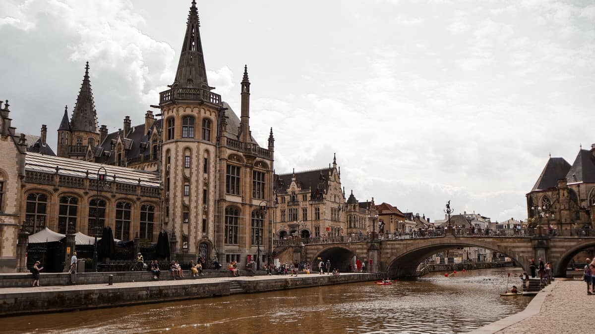 Classic view of Graslei and Korenlei canal in Ghent with historic medieval buildings, St. Michael’s Bridge, and people walking along the waterfront on a cloudy day.