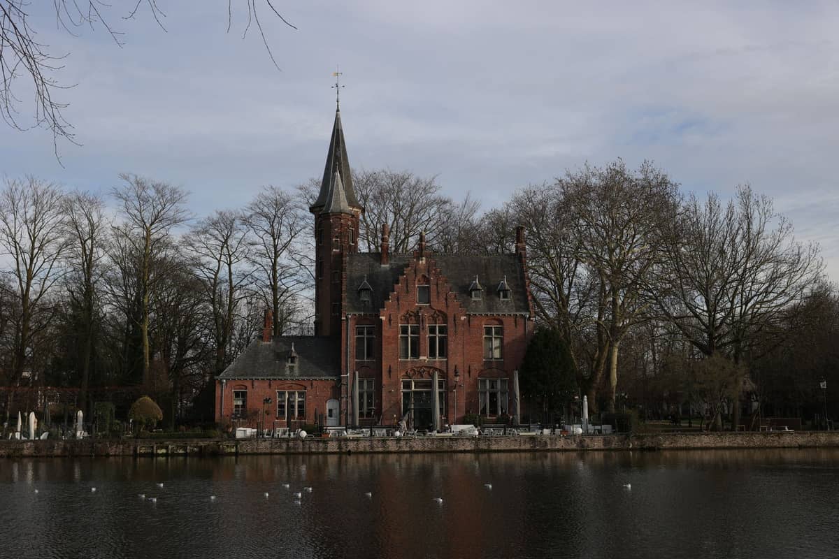 View across a peaceful pond to a distinctive red-brick, castle-like building surrounded by trees in a Ghent park setting.