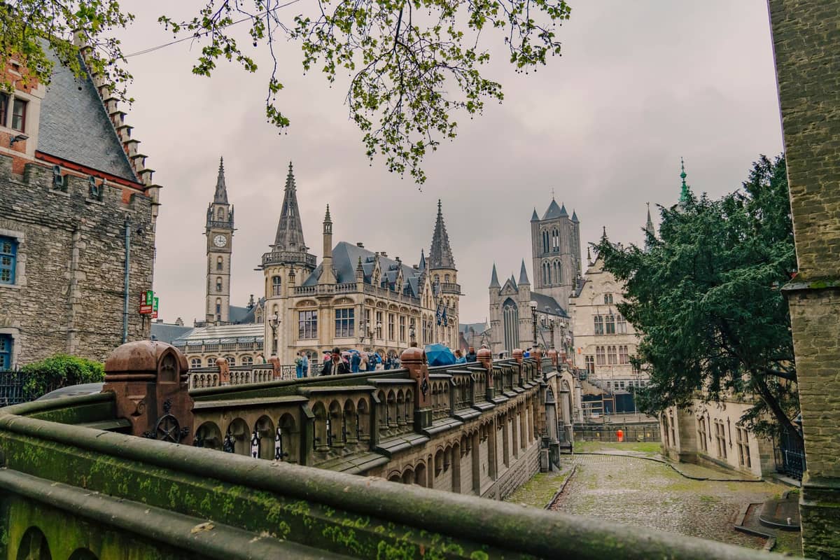 Classic panoramic view from St Michael's Bridge in Ghent showing the three medieval towers: St Bavo's Cathedral, the Belfry, and St Nicholas' Church under a cloudy sky.