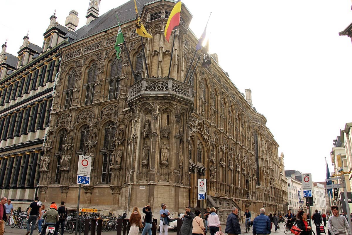 The ornate Gothic and Renaissance architecture of Ghent Town Hall with statues, flags, and people walking and cycling in the city street below.
