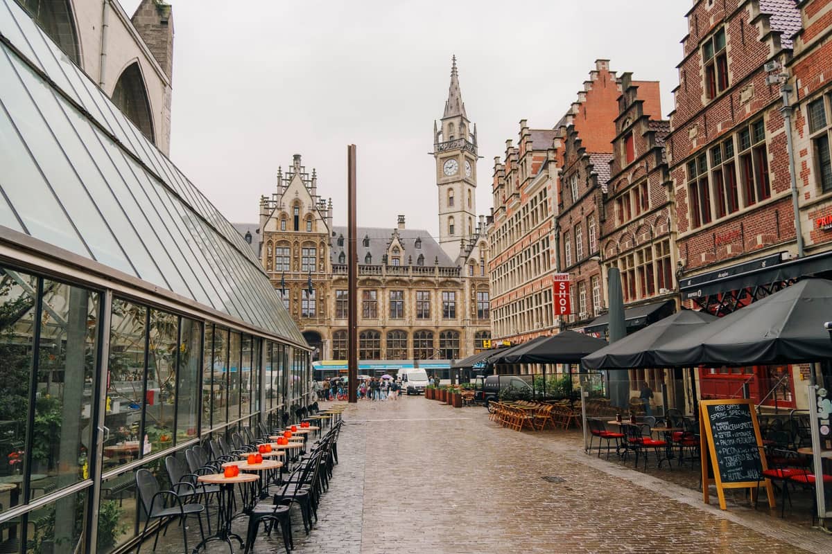 Café-lined street in Ghent with outdoor seating, colorful tables, and view of historic Flemish architecture and a clock tower in the background.