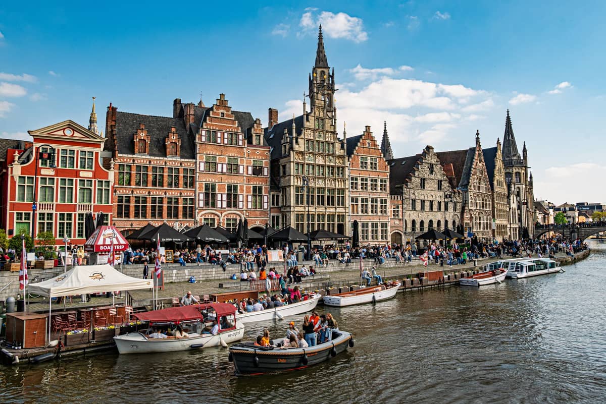 Vibrant and bustling canal scene in Ghent with historic Flemish buildings, outdoor cafes, and boats moored along the Graslei and Korenlei under a blue sky.