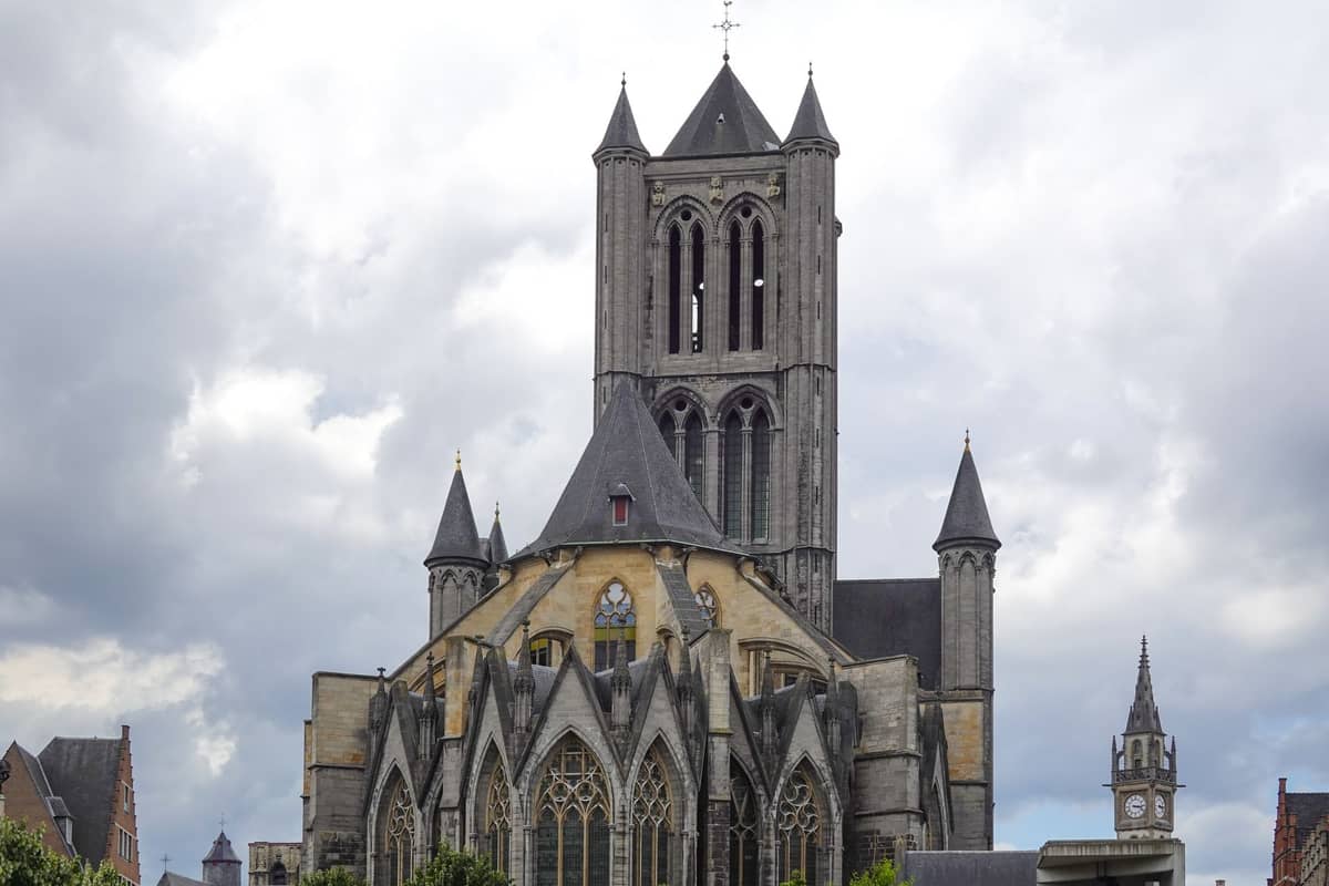 Exterior view of Saint Nicholas' Church in Ghent, showing gothic spires and large stained glass windows under a cloudy sky.