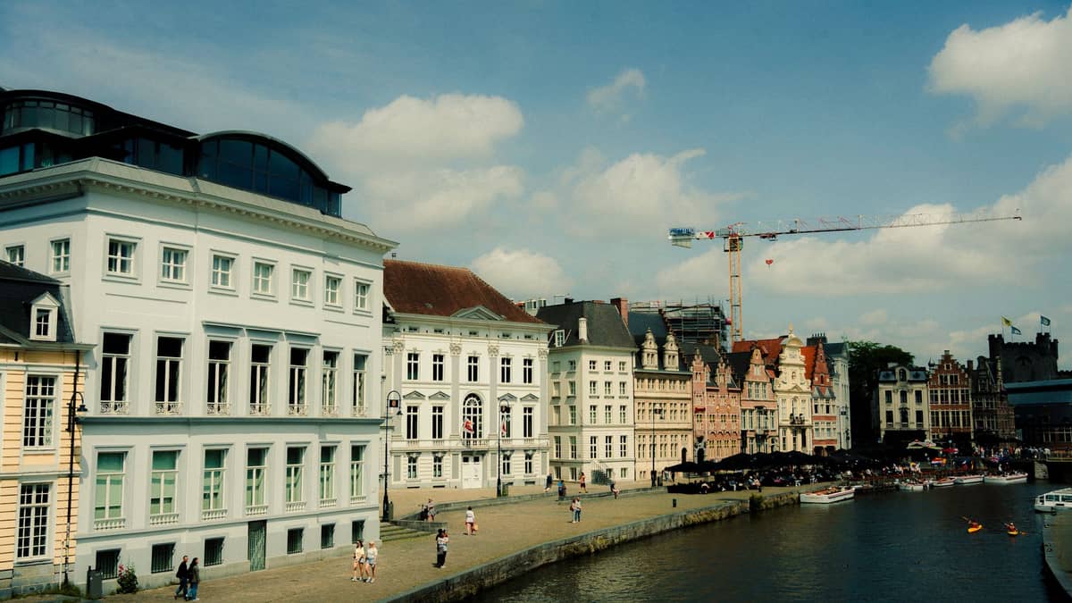 Daytime view of Ghent's Graslei canal lined with historic Flemish buildings, people strolling along the waterfront, and boat tours visible on the water.