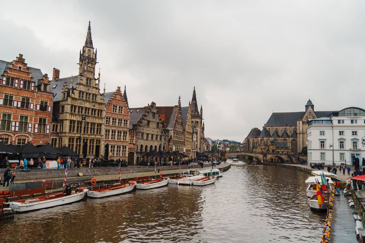 Wide view of Ghent’s main canal lined with historic guild houses, Gothic buildings, and several docked canal tour boats under a cloudy sky.