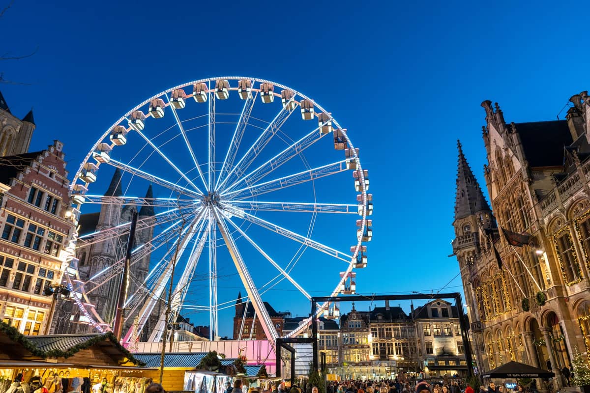 Large ferris wheel and festive market set up in Ghent's historic city center at dusk, with illuminated buildings and a lively crowd.