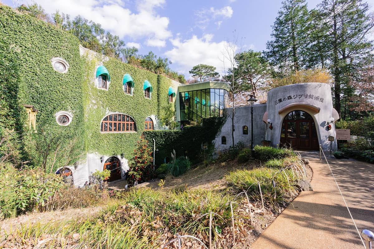 Wide view of the Ghibli Museum, Mitaka’s unique architecture, with lush ivy covering the exterior, whimsical windows, and the main entrance under a bright sky.