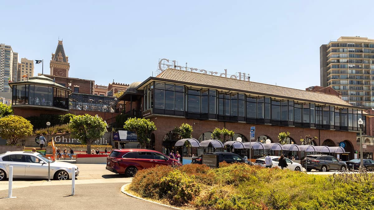 Vista diurna y amplia de los históricos edificios de ladrillo de Ghirardelli Square de la década de 1890, su famoso letrero en el techo y la entrada a la plaza, con personas, autos y jardines en primer plano.