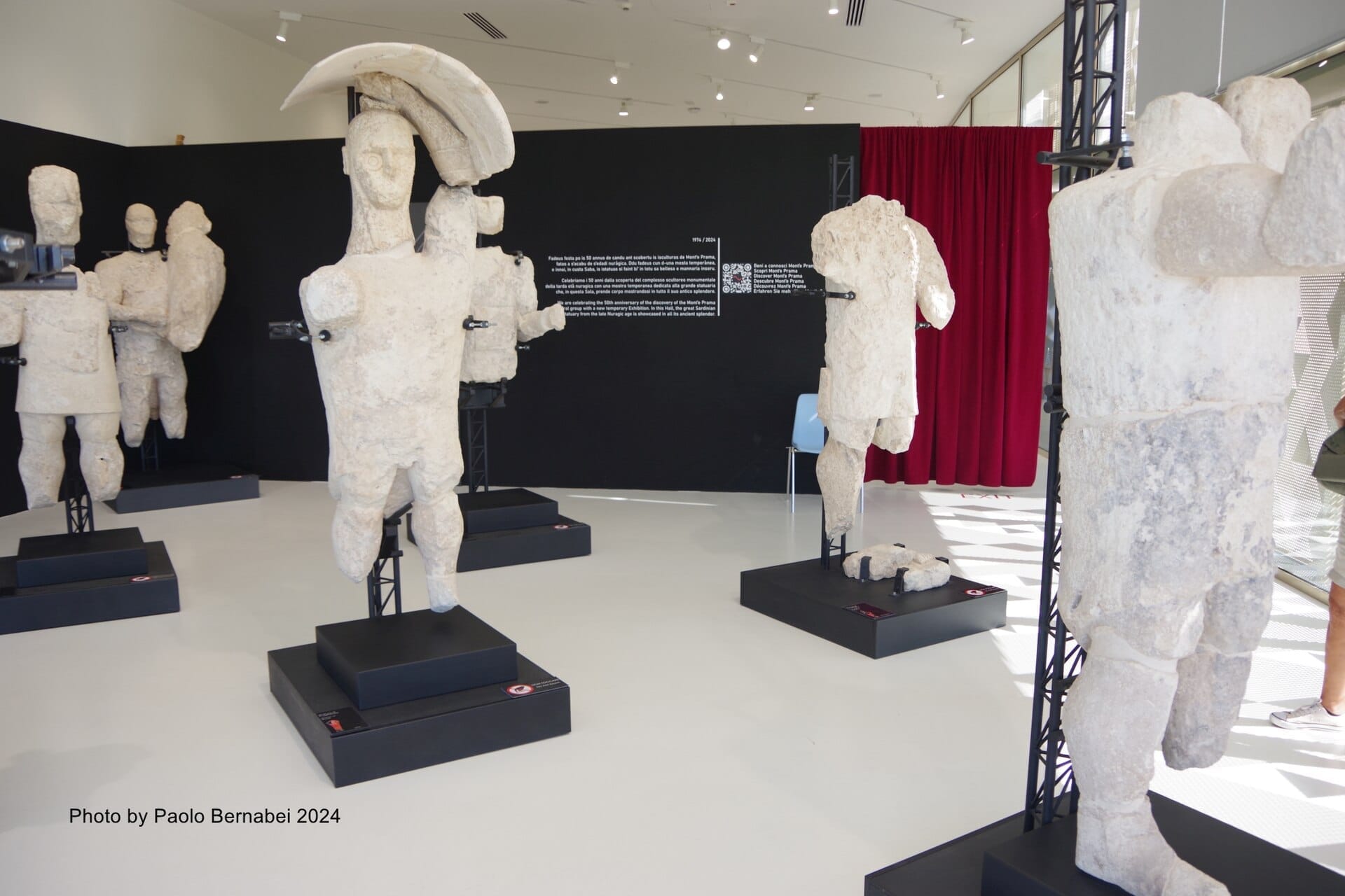 Mehrere antike Steinfiguren der Giganten von Mont'e Prama sind auf schwarzen Sockeln in einer hellen, modernen Ausstellungshalle ausgestellt.