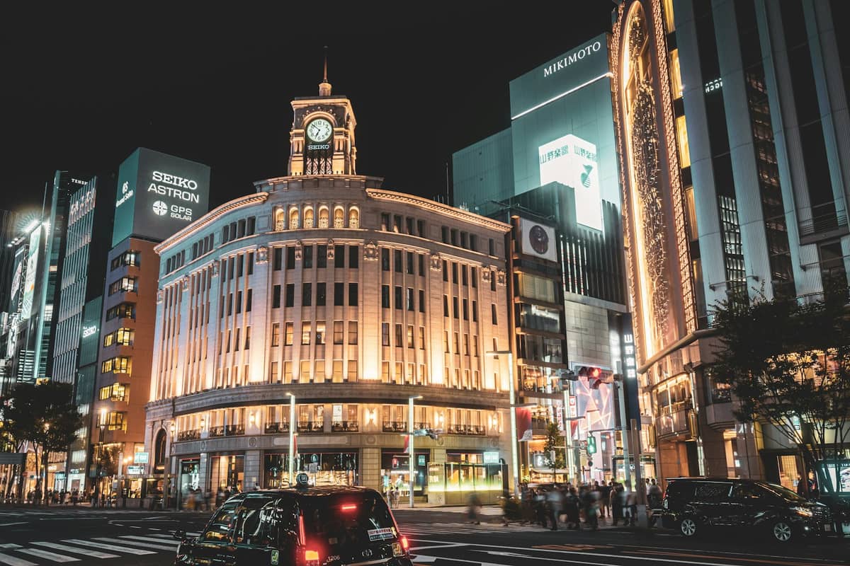 Night view of the iconic Wako building and shopping street in Ginza, Tokyo, with people, cars, and luxury storefronts illuminated.