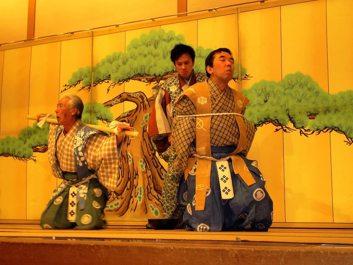 Three performers in traditional Japanese costumes act on stage at Gion Corner, with a painted pine tree backdrop and warm lighting.