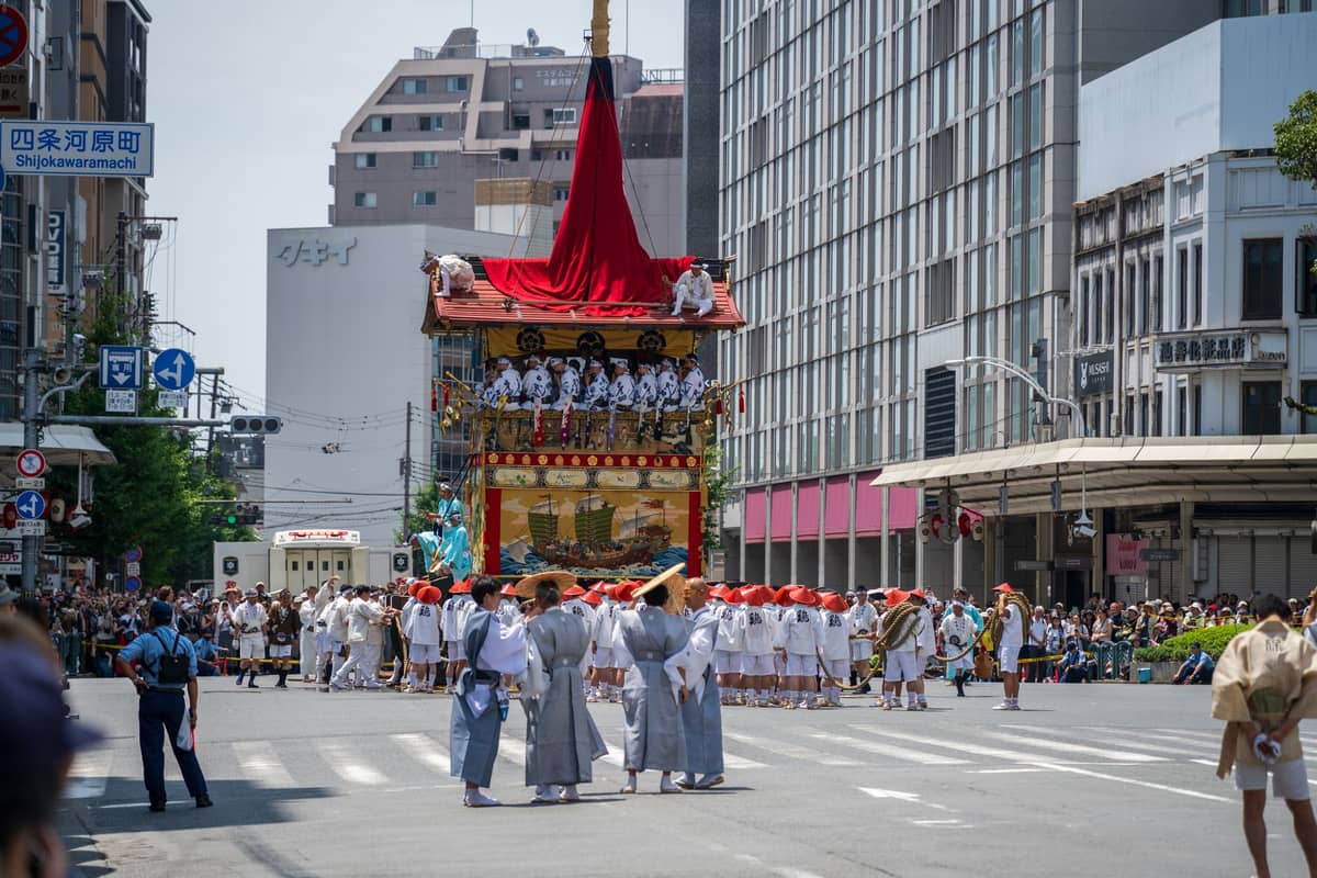 Crowds line a major Kyoto street as a grand Gion Matsuri float parades past, with clear signage for Shijo-Karasuma intersection.