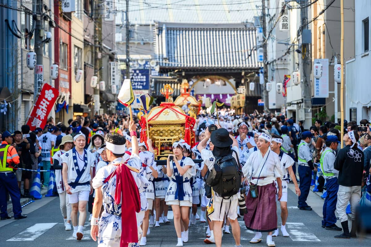 Large group in traditional festival attire carries a decorated mikoshi through crowded city streets, with Kyoto-style architecture visible.