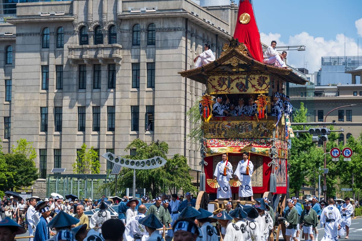 A large, ornate Gion Matsuri float being pulled along a city street by festival participants in traditional attire, surrounded by crowds and modern buildings in Kyoto.