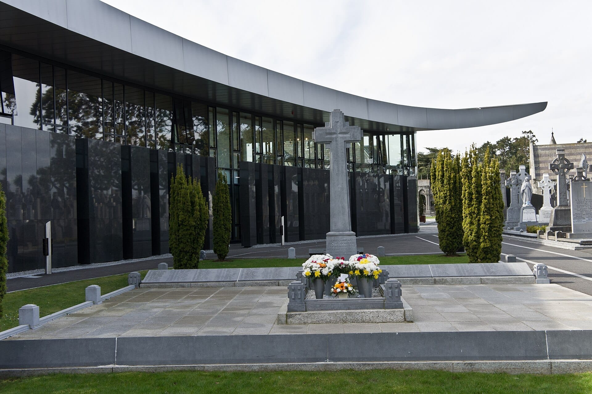 Moderno edificio in vetro del Glasnevin Cemetery Museum con monumento a croce in pietra, composizioni floreali e verde curato sotto un cielo nuvoloso.